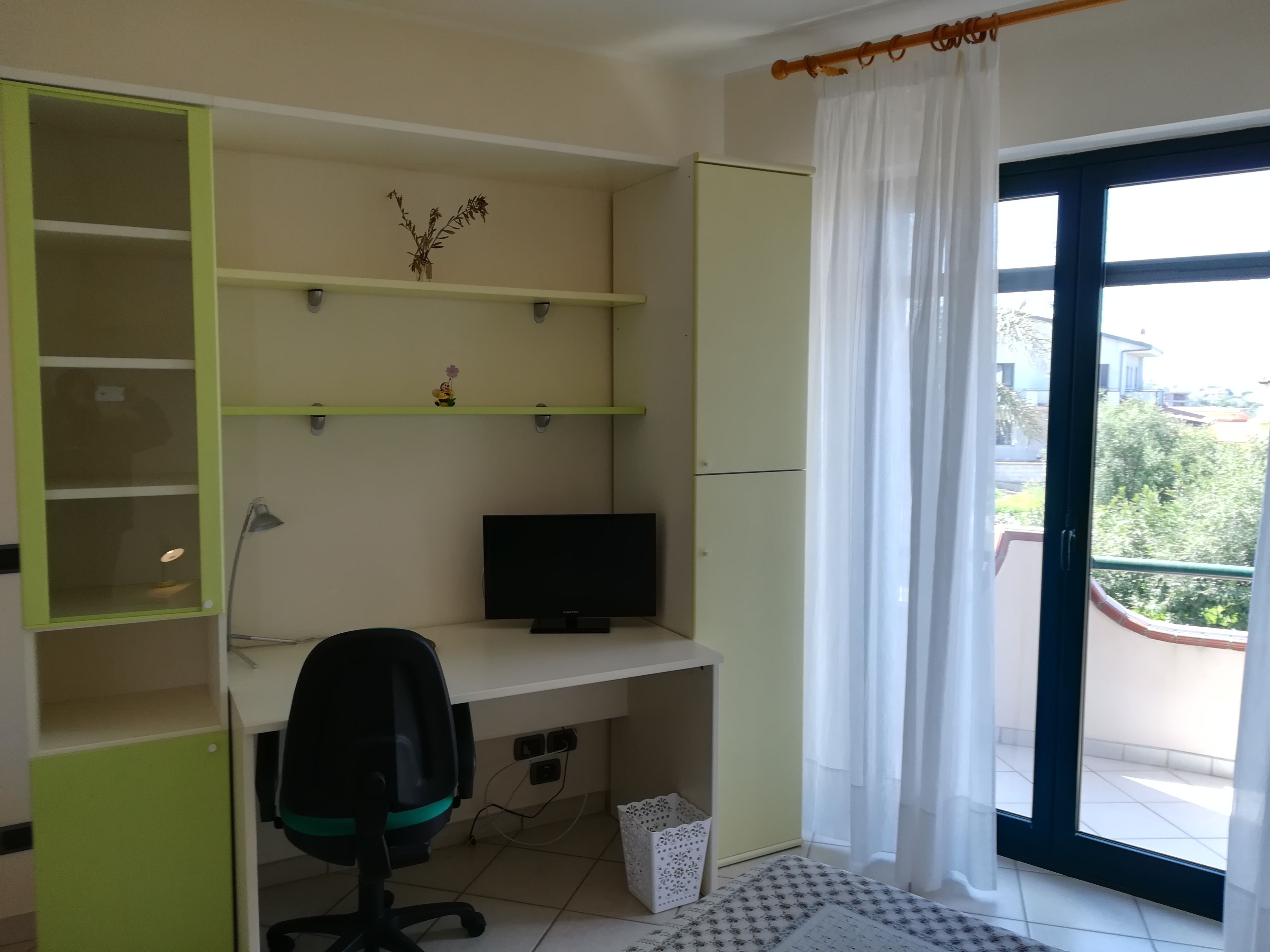 Ein Zimmer mit Schreibtisch, Stuhl, Regalen und einem Fenster mit Vorhängen.