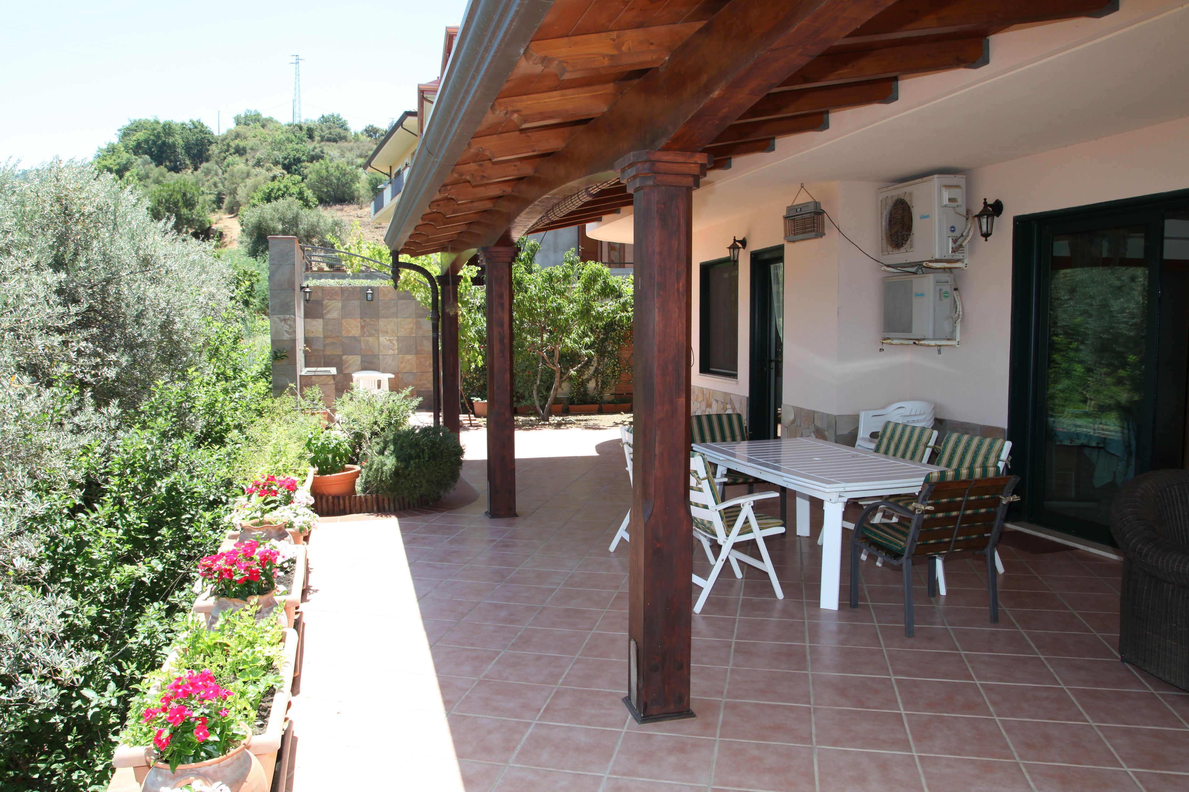 Een terras buiten met een houten pergola, een tafel met stoelen en plantenpotten.