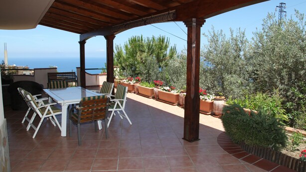 Un patio al aire libre con una mesa y sillas, rodeado de plantas en macetas y vista al mar.