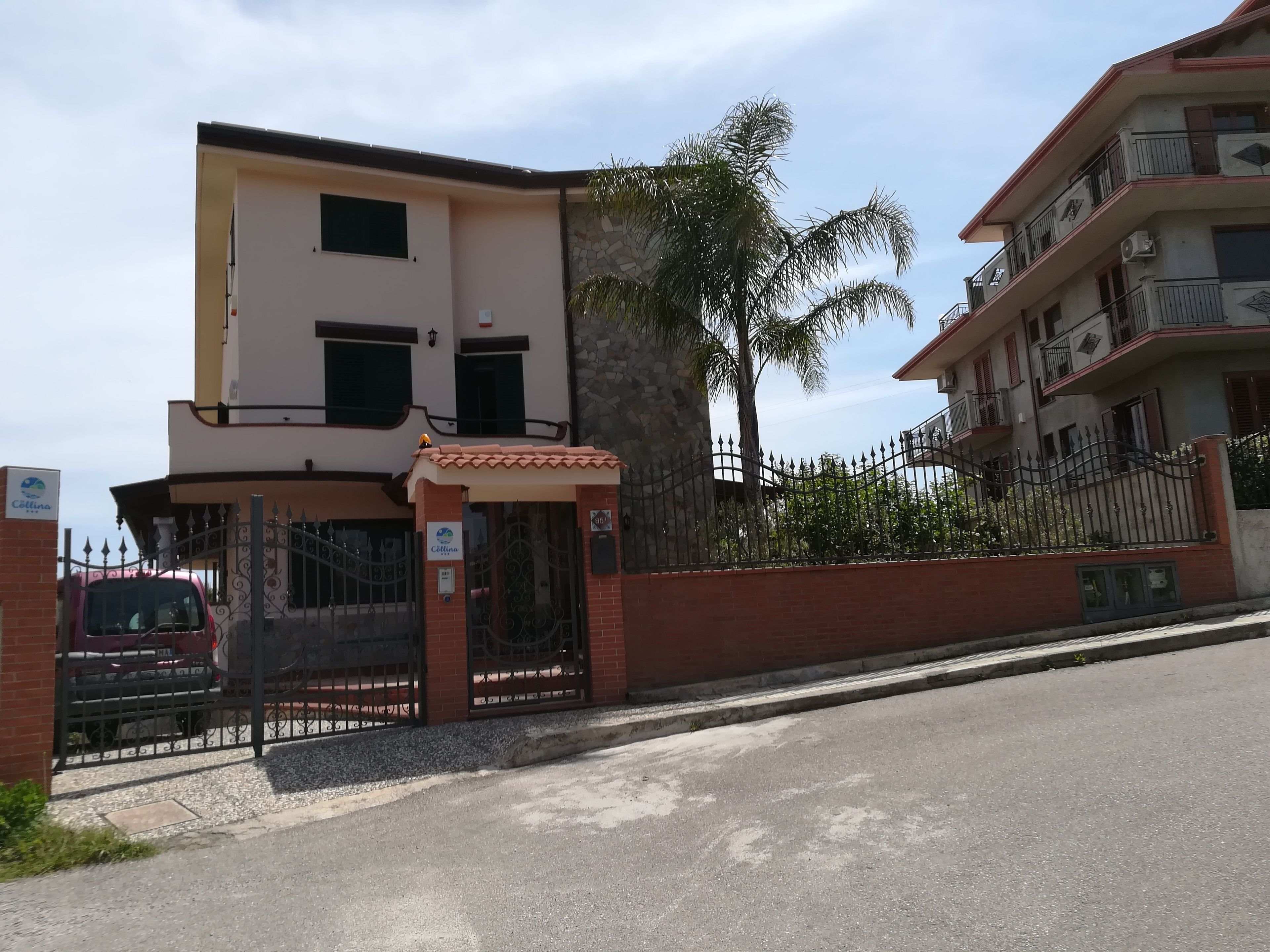 A multi-story residential building with a gated entrance, a red car parked outside, and a palm tree in the background.