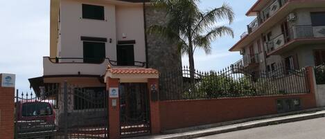 A multi-story residential building with a gated entrance, a red car parked outside, and a palm tree in the background.