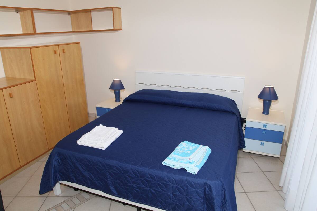 A neatly made bed with a blue bedspread, two bedside tables with lamps, a wooden wardrobe, and a white headboard.