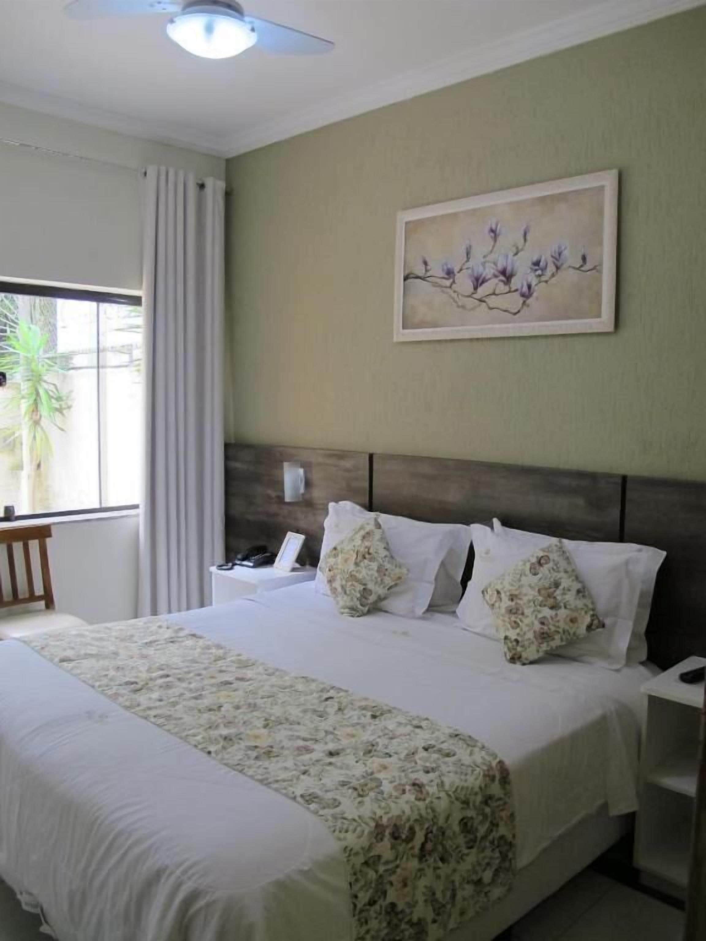 Una habitación de hotel con una cama grande, cabecera, mesita de noche con teléfono y ventana con cortinas.