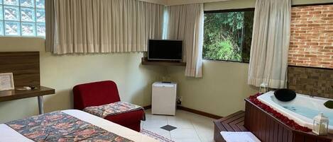 Habitación de hotel con dos camas, televisor, refrigerador pequeño, una silla roja, bañera con pétalos de rosa y ventana con cortinas.