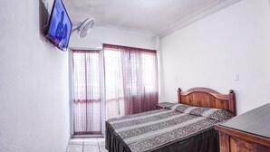 Un dormitori amb llit, televisió penjada a la paret, ventilador de sostre i finestra amb cortines.