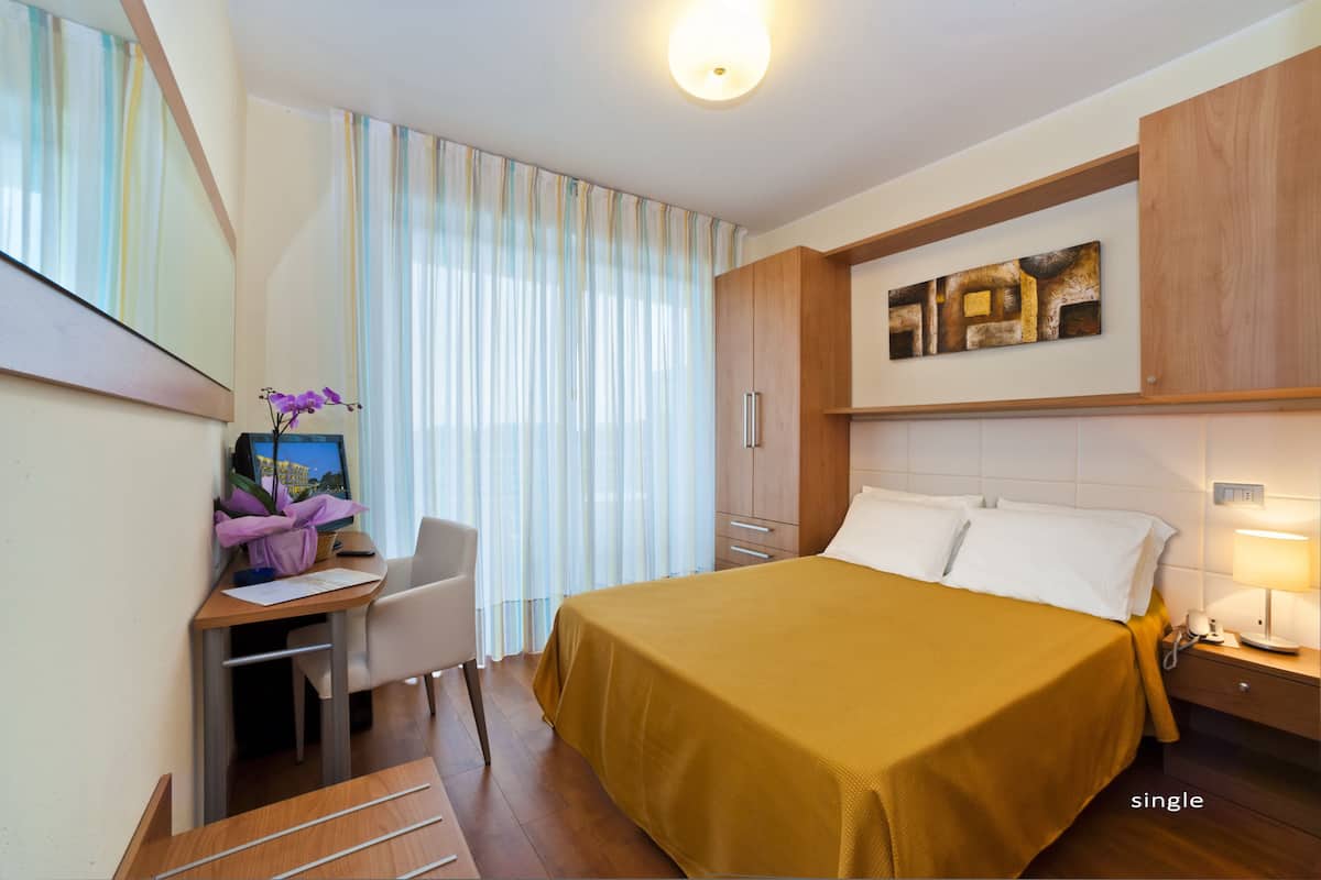 Een hotelkamer met een bed, een bureau met een computer, een stoel, een nachtkastje en een raam met gordijnen.