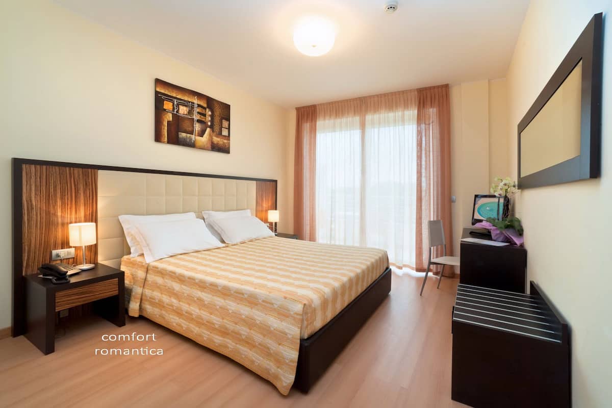Een hotelkamer met een groot bed, een nachtkastje met een lamp, een bureau met een stoel en een grote spiegel aan de muur.