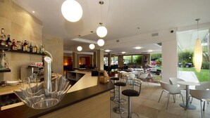 A modern bar area with a clear glass bar counter, multiple hanging lights, and a view of the outdoor seating area. - Hotel San Marco (San Michele al Tagliamento)