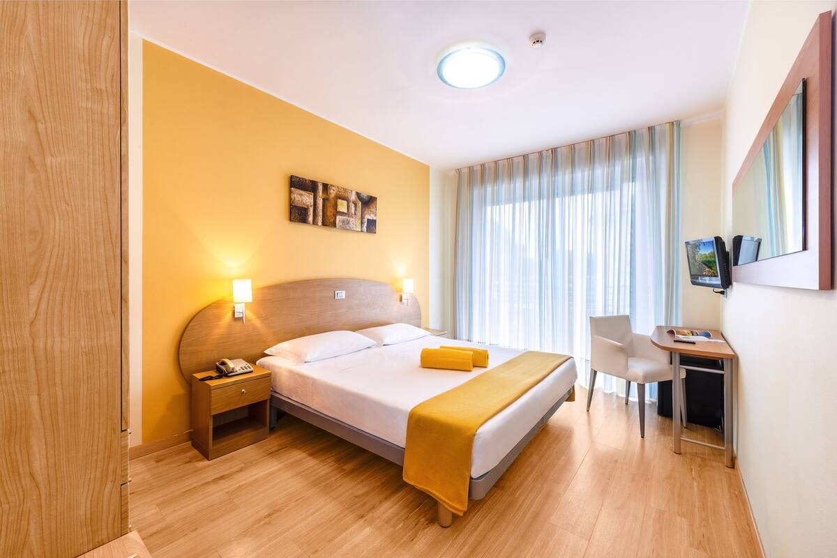 Hotelkamer met een bed, nachtkastje, bureau, stoel en een groot raam met gordijnen.