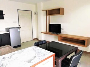 Un espacio habitable compacto con zona de cocina, una mesa de comedor con sillas, un televisor montado en un estante de madera y un refrigerador.