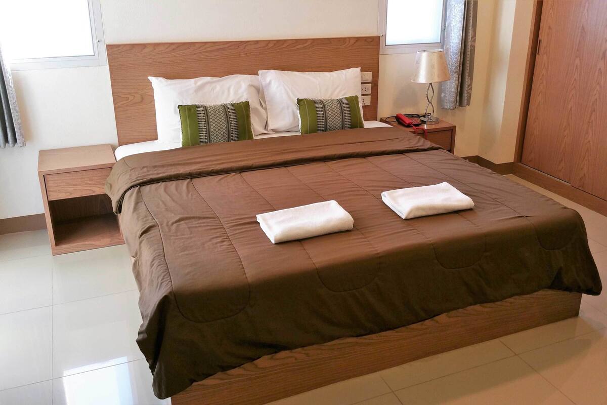 A neatly made bed with a brown bedspread and two white towels, a wooden headboard, and a bedside table with a lamp.