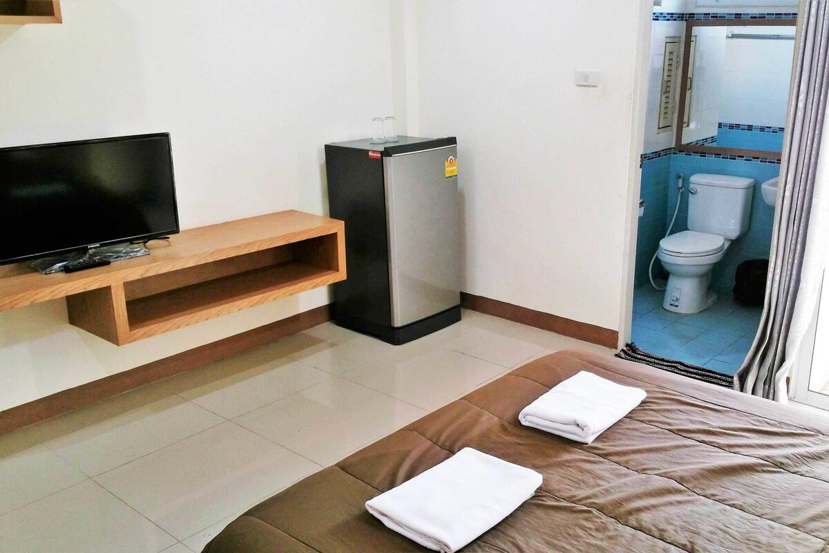 A hotel room with a bed, a TV on a wooden shelf, a refrigerator, and a view of the bathroom.