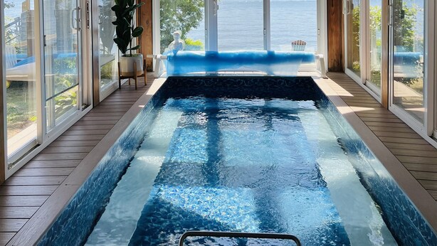 Une piscine à débordement en bois, dotée d’un ventilateur de plafond, de grandes fenêtres et offrant une vue sur le lac.