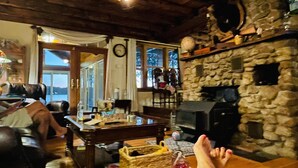 Un salon chaleureux avec une cheminée en pierre, un plafond en bois et une vue dégagée sur l’extérieur grâce à une grande fenêtre.