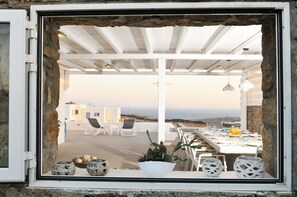 Une fenêtre ouverte donne sur une terrasse où l’on aperçoit une table à manger dressée pour deux personnes, des chaises blanches et un cactus en pot.