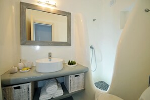 Une salle de bain avec un lavabo blanc, un miroir et une douche. On trouve des paniers sous le lavabo et des serviettes sur le sol.