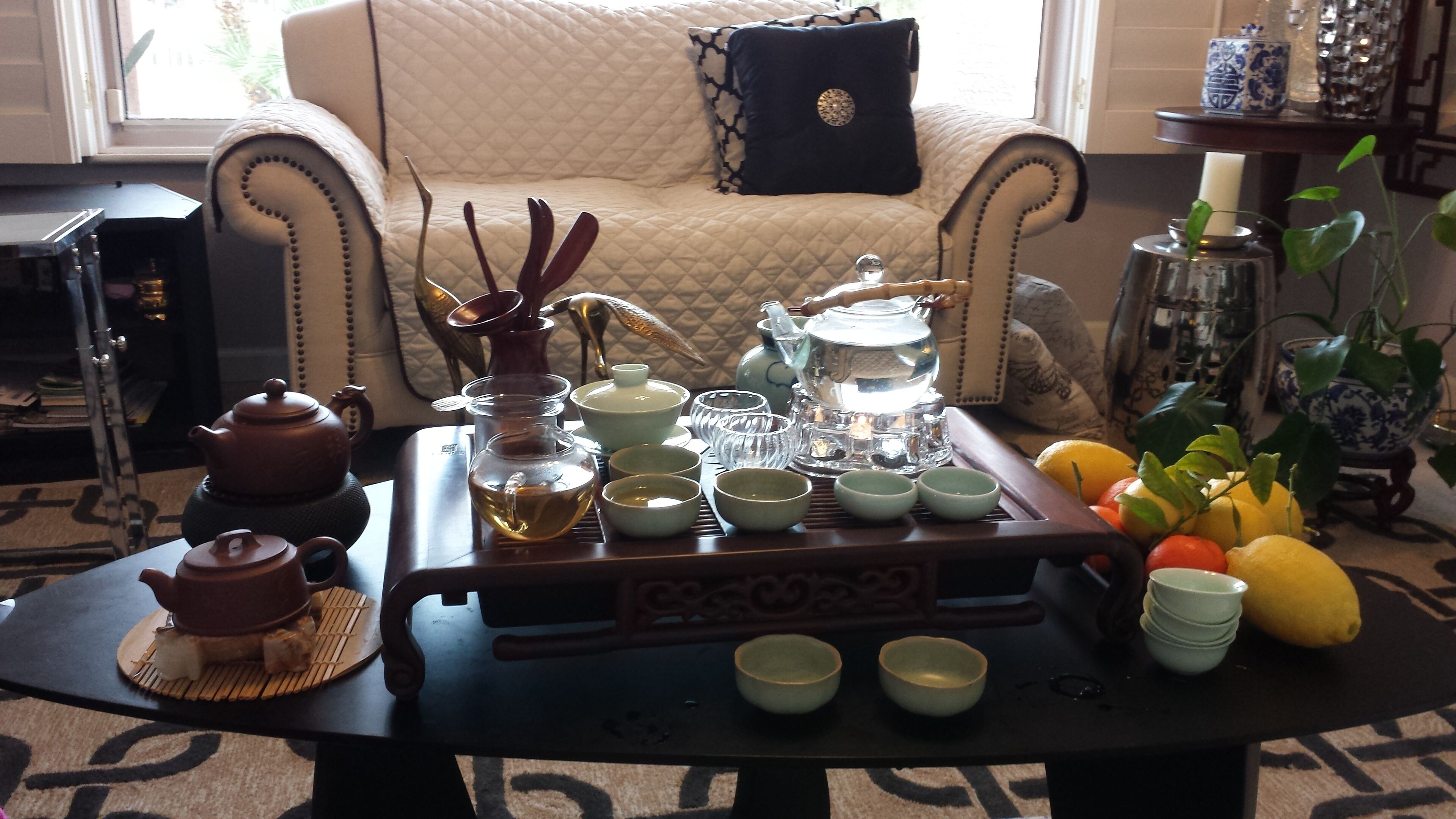 Un juego de té tradicional sobre una mesa, con un tapete estampado, un sofá con un cojín decorativo y una ventana que deja entrar luz natural.