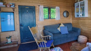 Interior acolhedor de uma cabana com uma porta azul, um sofá, uma cadeira de madeira e um pequeno armário com objetos decorativos.