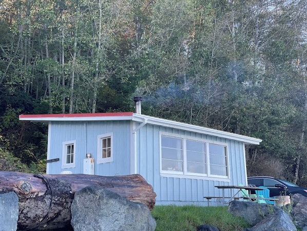 A small, light blue cabin with a red roof, surrounded by trees and rocks.