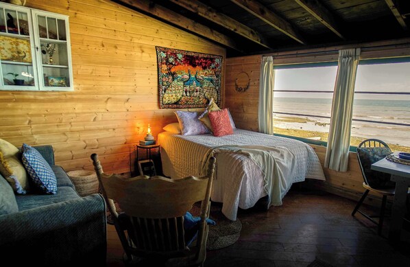 A cozy cabin room with a bed, two chairs, a table, and a view of the beach.