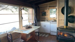 A cozy cabin interior with a wooden dining table, chairs, a kitchen area with a stove, and a wood-burning stove.