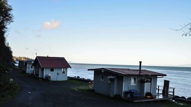 Una propiedad costera con dos cabañas pequeñas, una de techo rojo y la otra de techo gris, ambas con porches anexos y vistas al mar.
