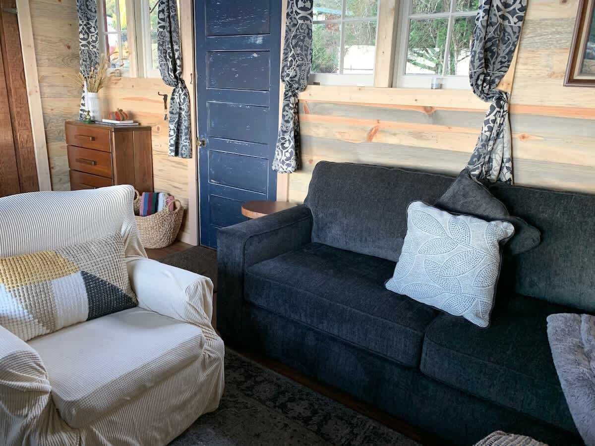 A cozy living room with a dark grey sofa, a white armchair with patterned cushions, and a wooden cabinet.