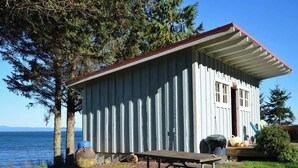 A small, single-story cabin with a red roof and white siding, located near a body of water with trees around.