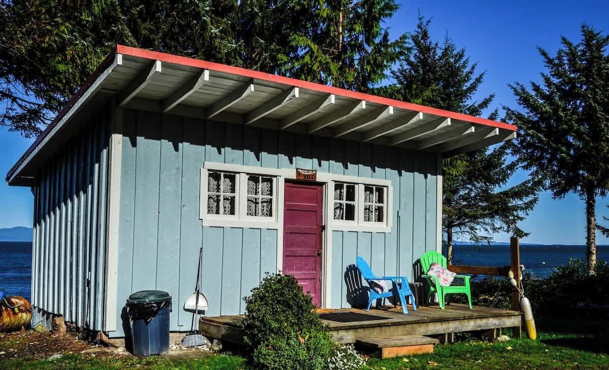 A small, single-story cabin with a red roof and a front door. There are two chairs outside and a trash can to the left.