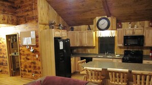 A wooden cabin interior with a kitchen featuring wooden cabinets, a black refrigerator, a microwave, and a clock. There is a wooden dining table with chairs and a purple throw on a sofa.