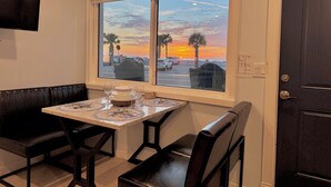 Un área de comedor con una mesa preparada para dos, un banco y vistas a la puesta de sol sobre un estacionamiento con palmeras.