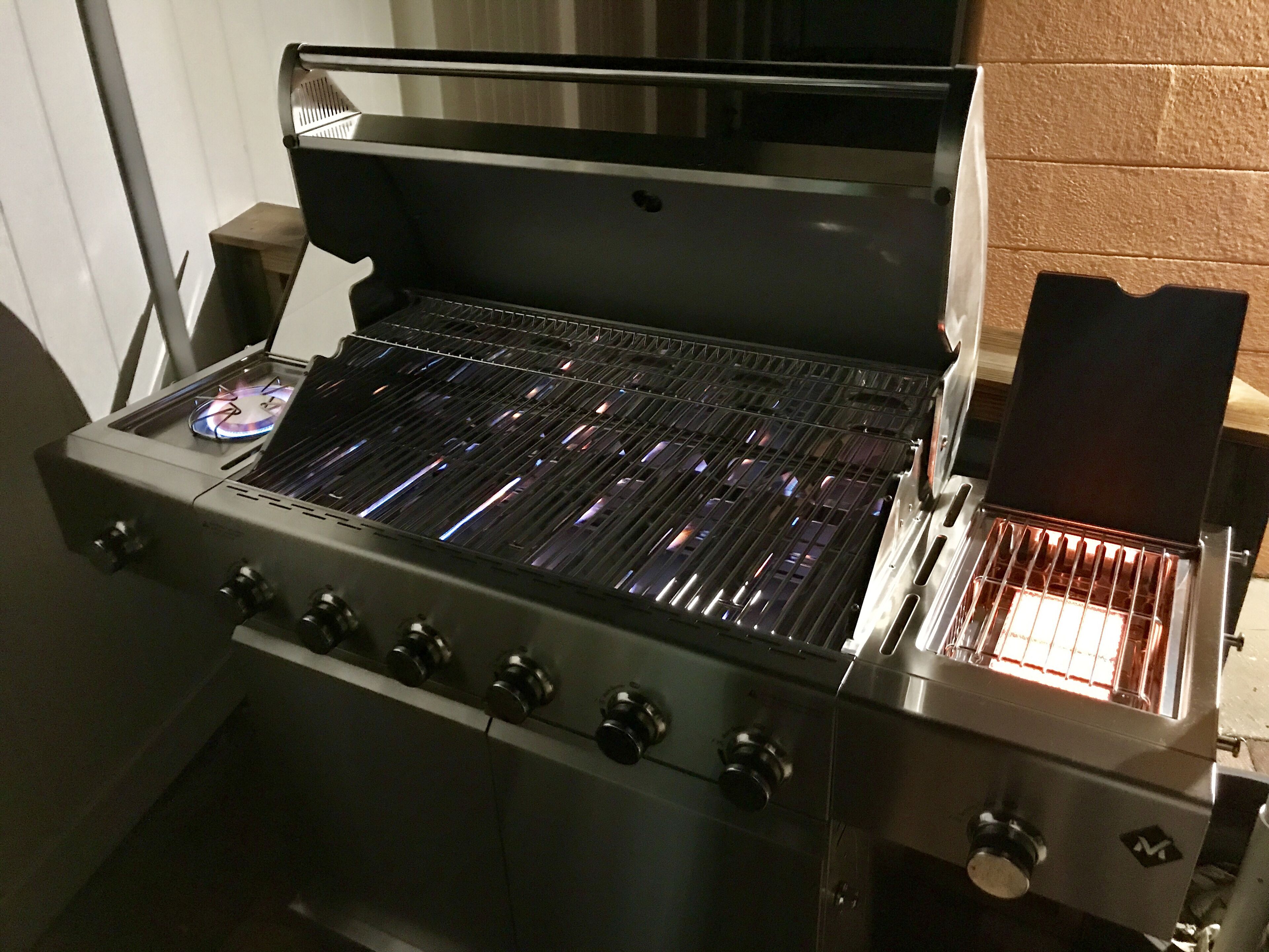 Una parrilla de gas portátil con acabado de acero inoxidable, una tapa de vidrio y un quemador lateral.