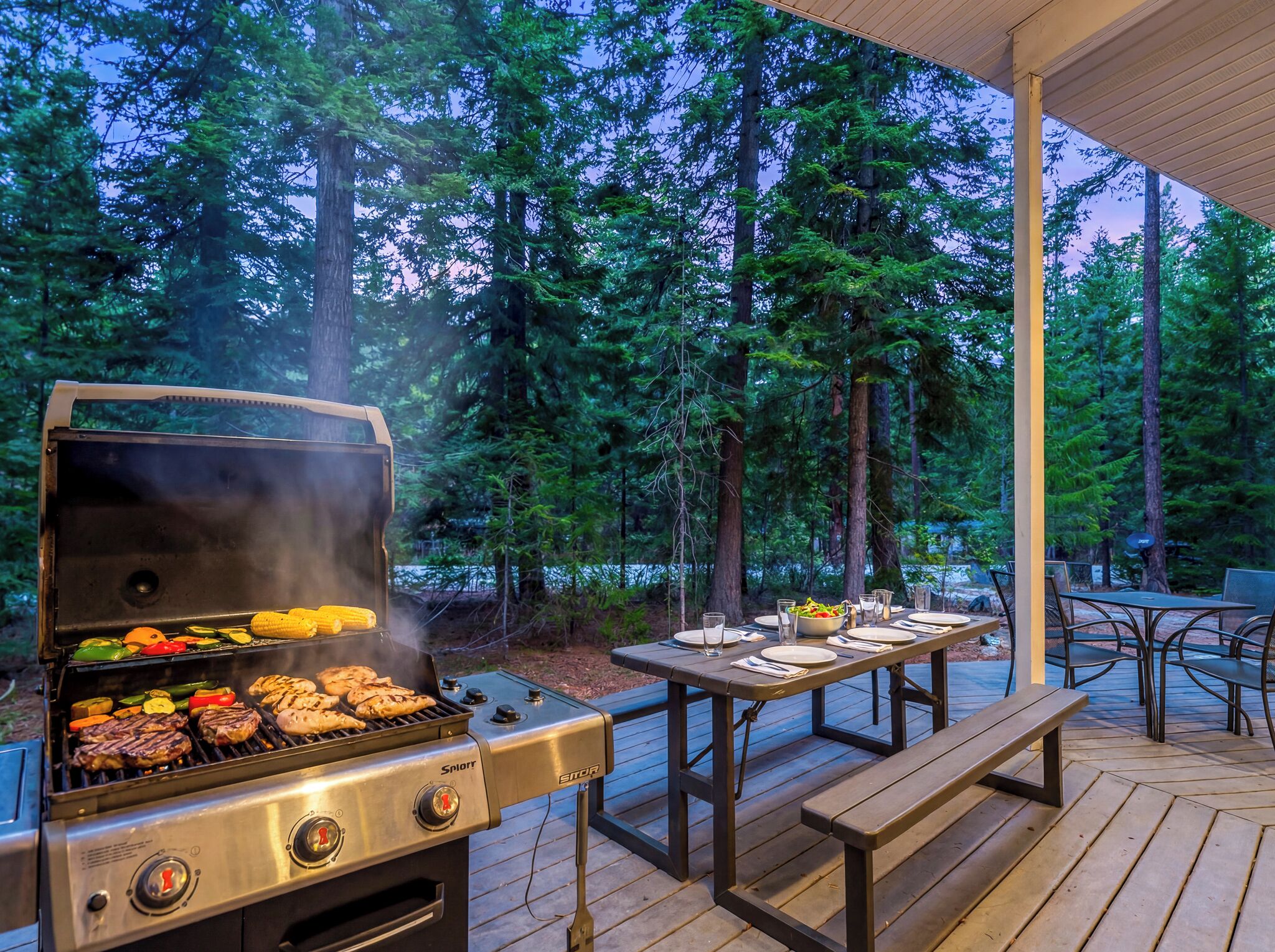 An outdoor grill with food, a covered dining area, and a forested backdrop.