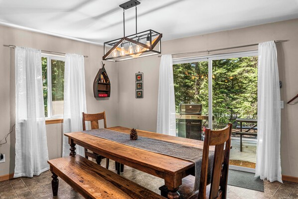 A dining area with a wooden table, chairs, a bench, a hanging light fixture, and a view through glass doors to an outdoor deck.