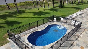 Ein Swimmingpool mit schwarzem Metallzaun, umgeben von einer Steinterrasse und einer Rasenfläche mit Palmen.