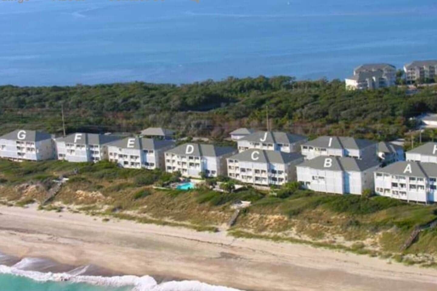 A coastal hotel with multiple buildings labeled with letters, a beach in the foreground, and a view of the ocean.