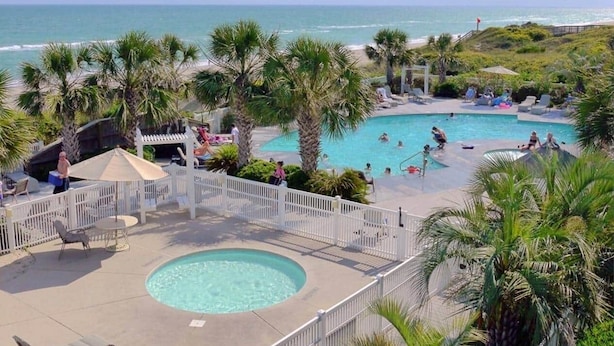 A pool area with a view of the ocean, surrounded by palm trees and a white fence.