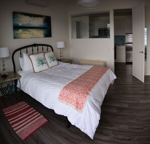 A bedroom with a bed, nightstands, a red and white striped rug, a painting, and a view of the kitchen.
