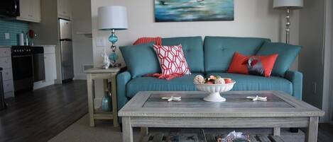 A living room with a teal sofa, a coffee table, and a kitchen in the background.
