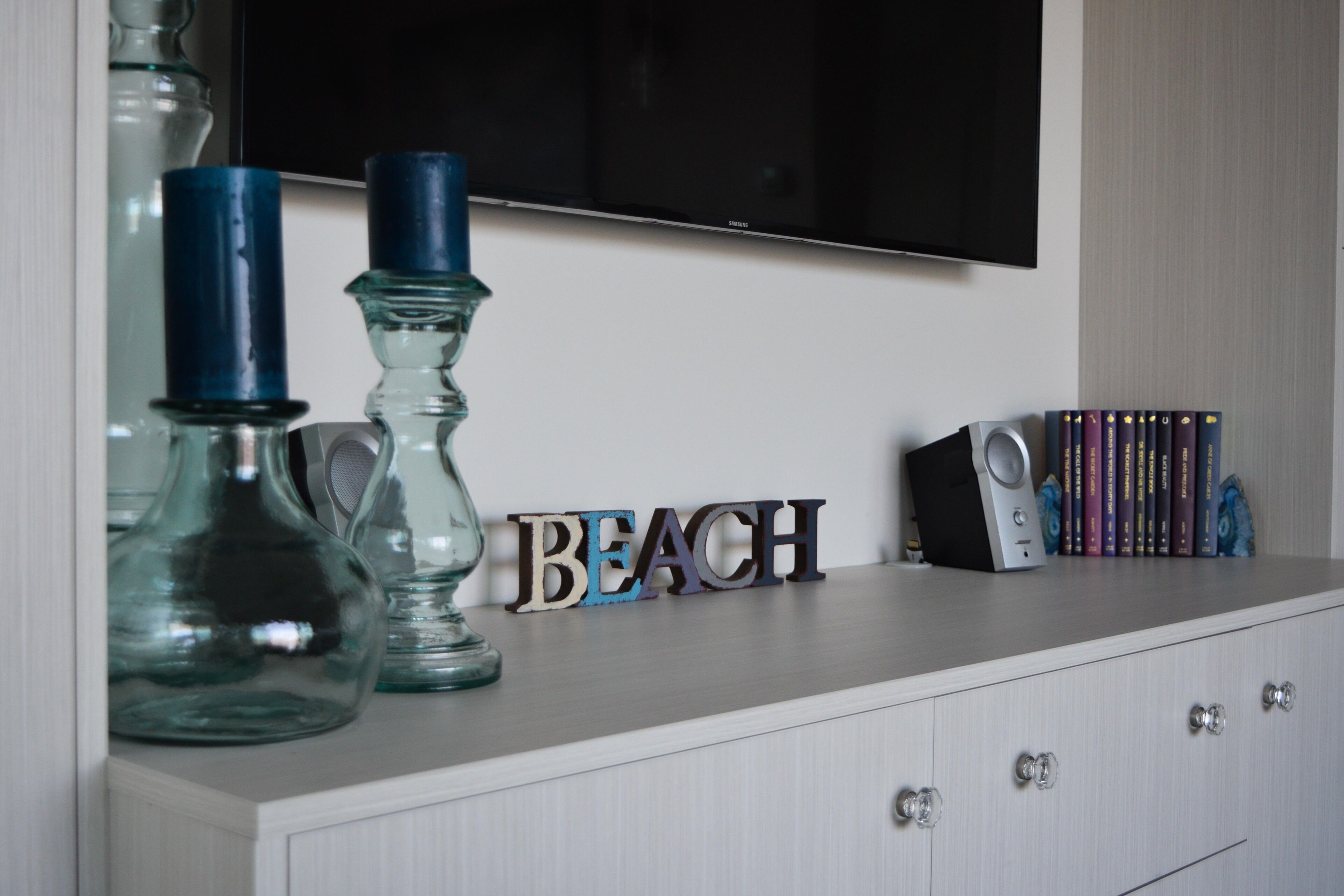Un meuble blanc sur lequel est fixé un téléviseur à écran plat, avec le mot décoratif « BEACH » inscrit sur le meuble, et une rangée de livres à droite.