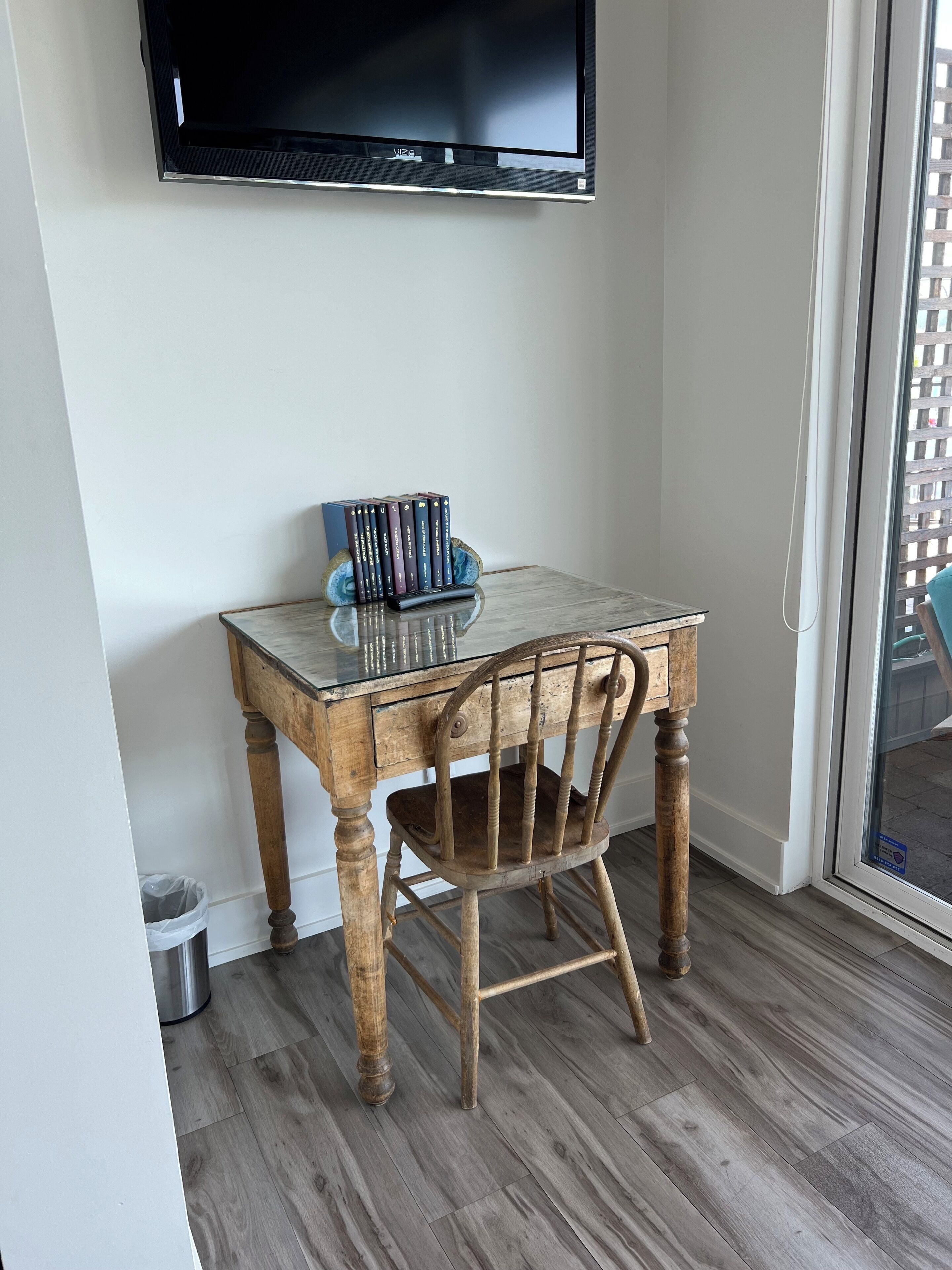 Une petite table en bois surmontée d’une plaque de verre, une chaise et une pile de livres. Un téléviseur à écran plat est fixé au mur au-dessus de la table.