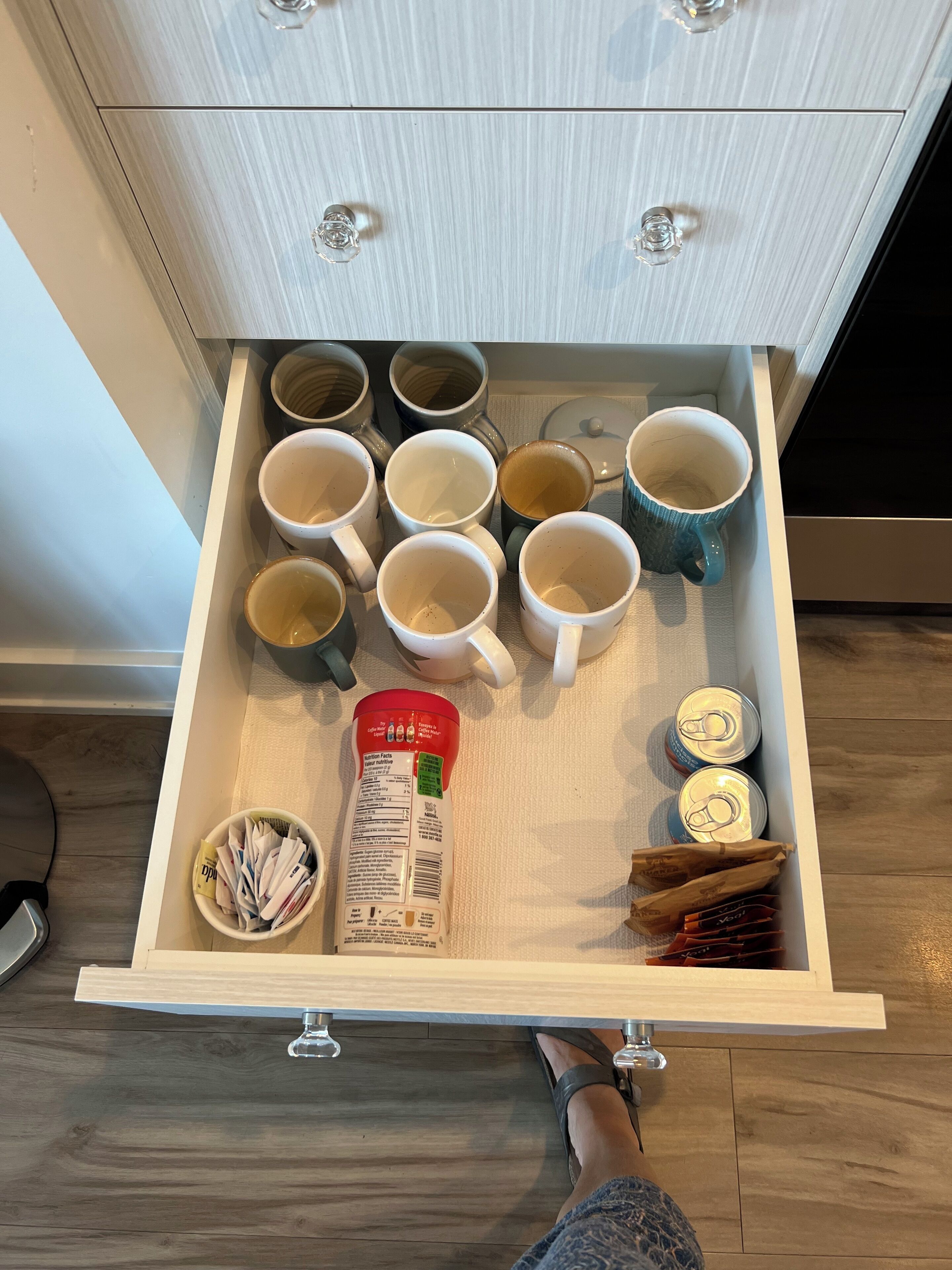 A drawer with multiple mugs, a bottle of coffee creamer, and a few cans.