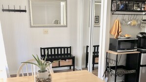 Ruokailutilassa on puinen pöytä, valkoiset tuolit, musta penkki ja ruukkukasvi. Keittiöalueella on kodinkoneita ja keittiövälineitä.