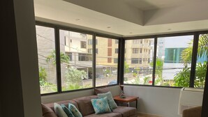 Un salón amplio con un sofá marrón, varios cojines, una mesita y ventanas grandes que ofrecen vistas a edificios y zonas verdes.