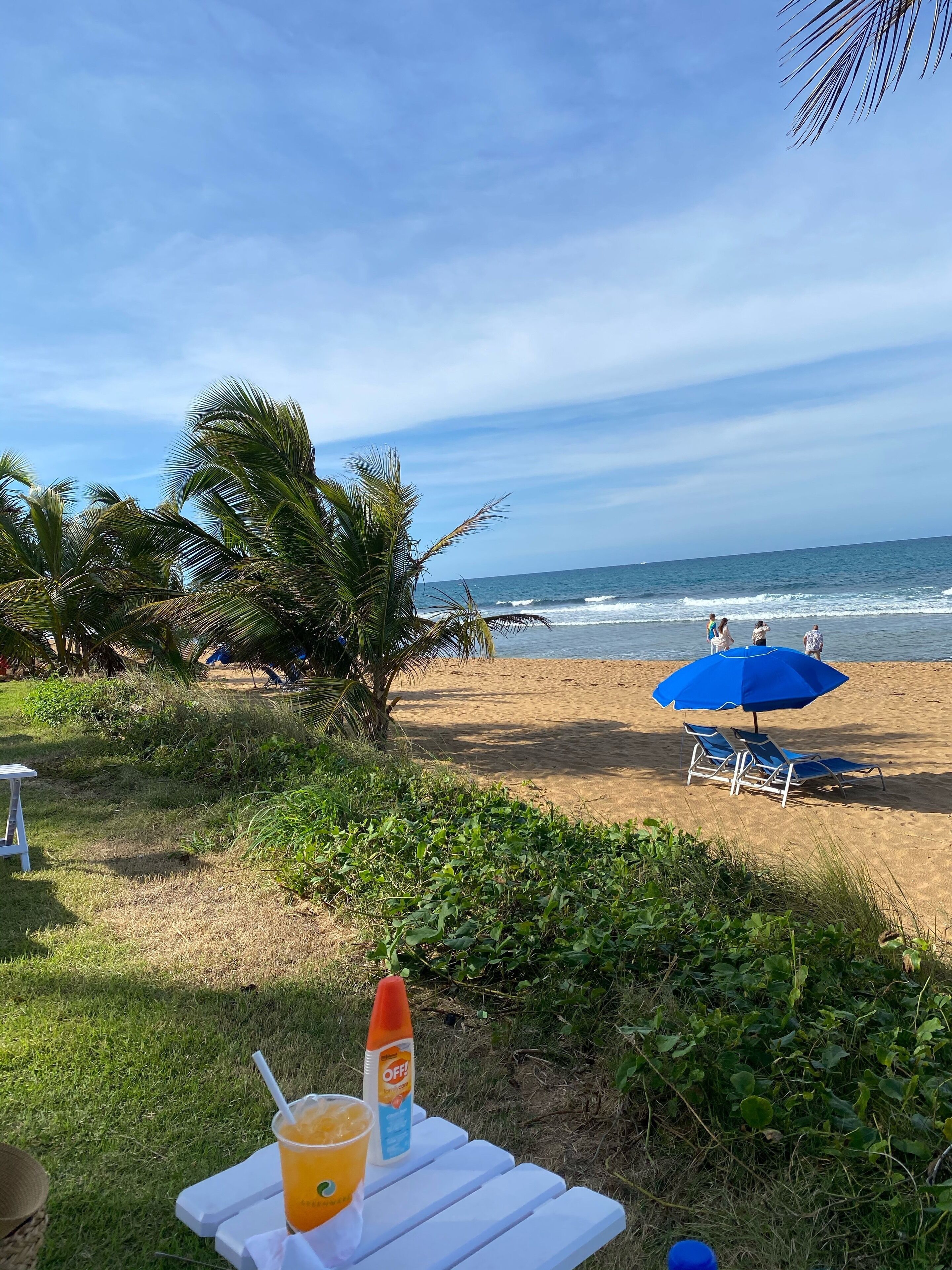 A beachside table with a drink and sunscreen, a blue umbrella, and a chair.