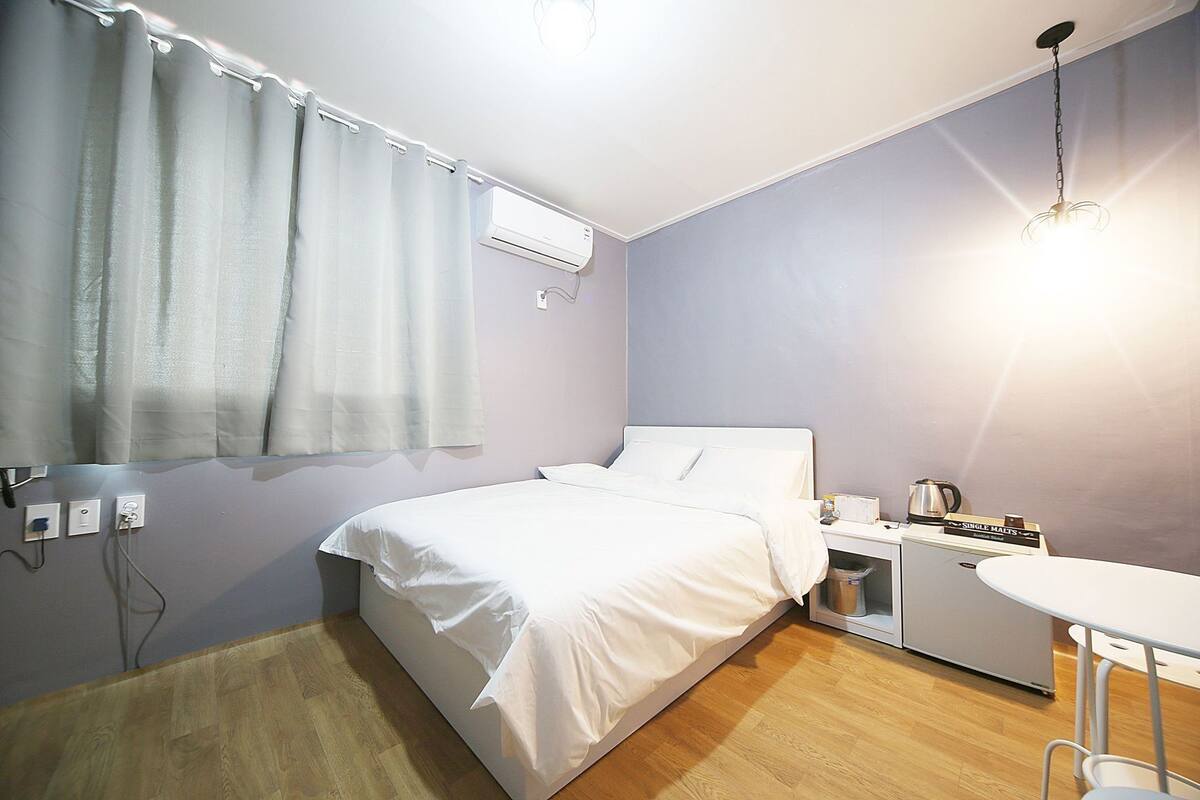 A bedroom with a bed, a bedside table with a kettle, a small table, and a wall-mounted air conditioner.