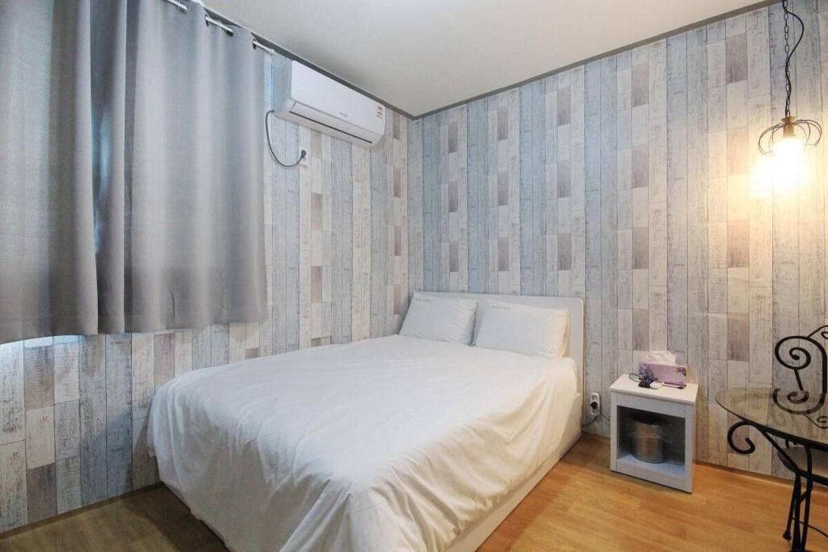 A bedroom with a bed, a nightstand, a table, and an air conditioning unit.