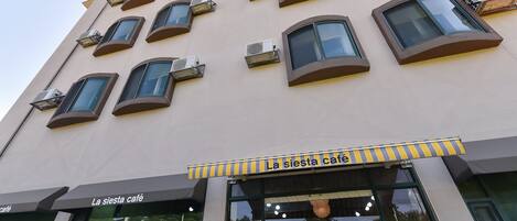 Un edificio con varios balcones y unidades de aire acondicionado. La planta baja tiene una cafetería llamada "La siesta café" con ventanas grandes y terraza.