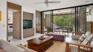 Un salon moderne doté d’un téléviseur à écran plat, d’un ventilateur de plafond et d’une grande porte-fenêtre coulissante donnant sur un balcon avec vue sur les arbres.