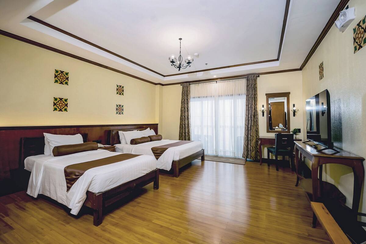 A hotel room with two beds, a desk, a television, and a chandelier.